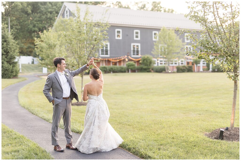 groom twirls his bride under his left arm along pathway outdoors with gray barn behind them at historic ashland