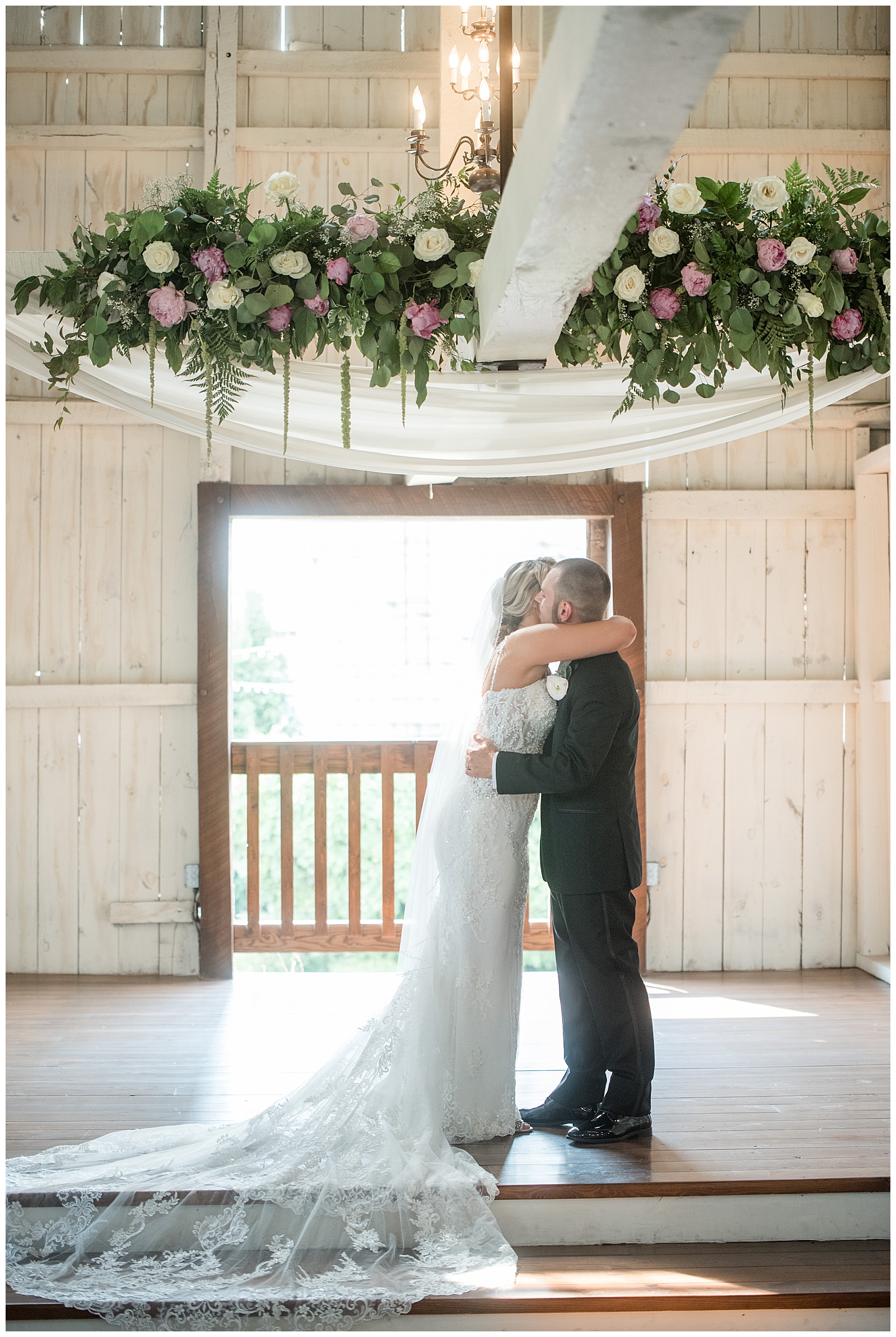 bride and groom hugging and kissing during barn wedding ceremony by large floral display with doorway open behind them at stoltzfus homestead