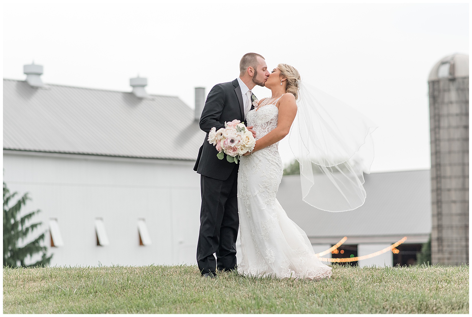 groom slightly dipping bride back as they kiss with beautiful white barn in background at stoltzfus homestead and gardens