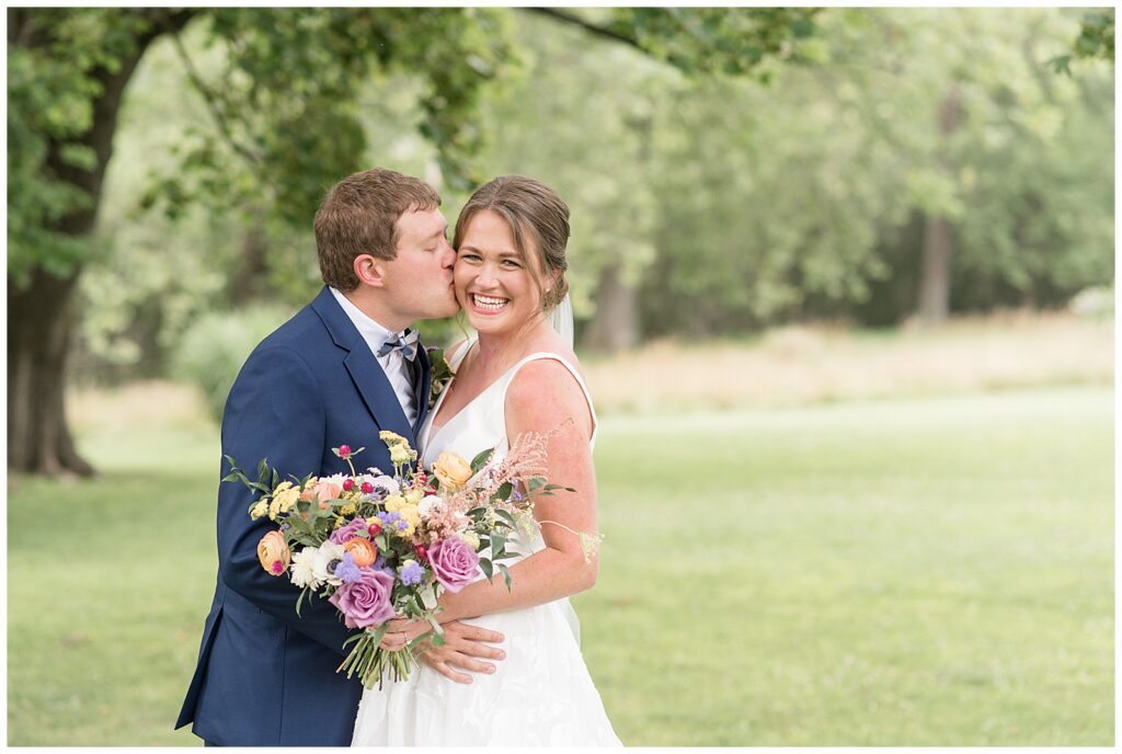 groom kissing bride on her right cheek as she smiles at camera holding bouquet outdoors at the inn at grace winery