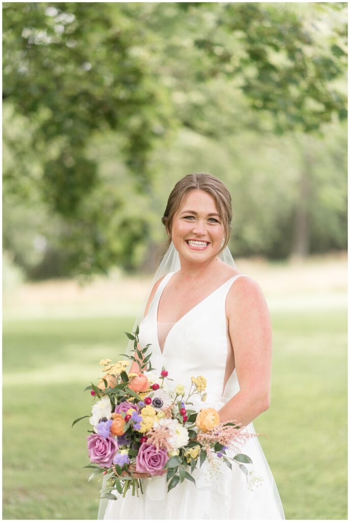 bride in white v-neck sleeveless gown holding colorful bouquet by trees at the inn at grace winery