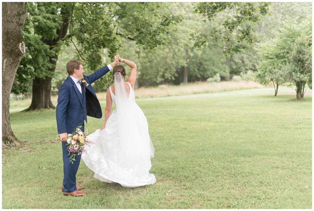 groom twirling bride under his left arm outdoors by large trees as he holds her bouquet in glen mills pa