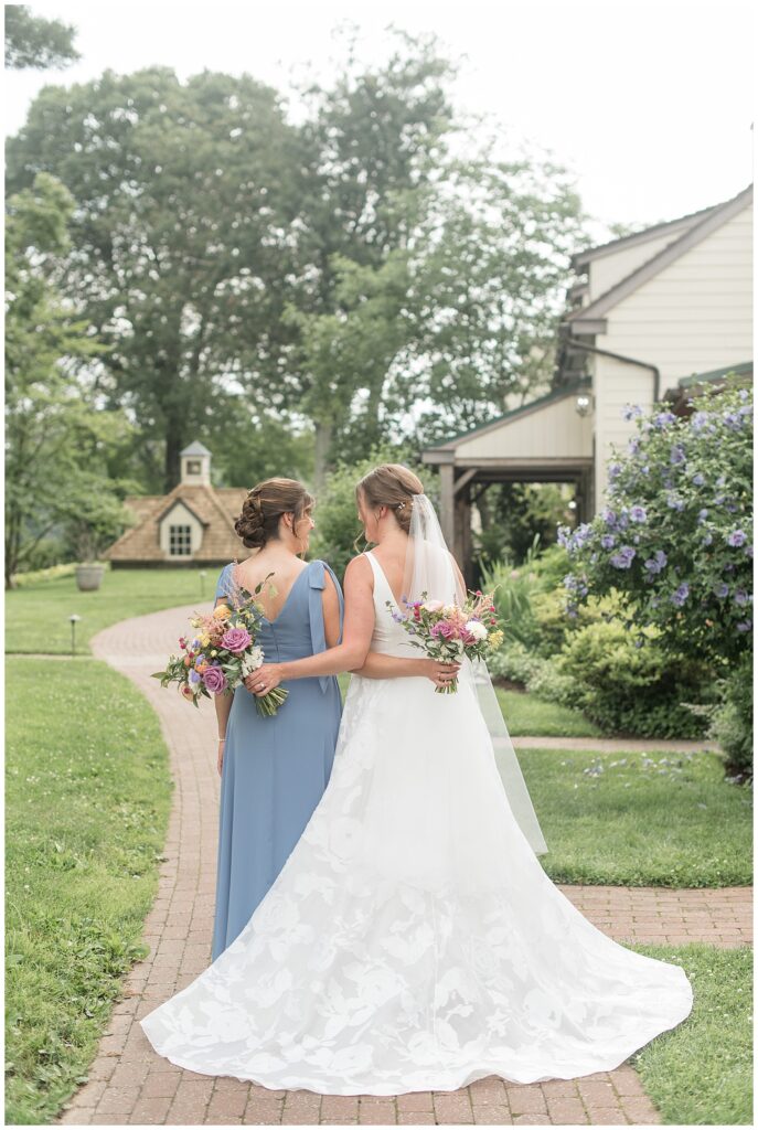 bride and her bridesmaid in pale blue gown with backs toward camera and arms around one another on pathway at the inn at grace winery