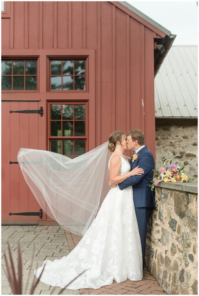 groom leaning against stone wall by red barn kissing his bride with her veil blowing behind her at the inn at grace winery