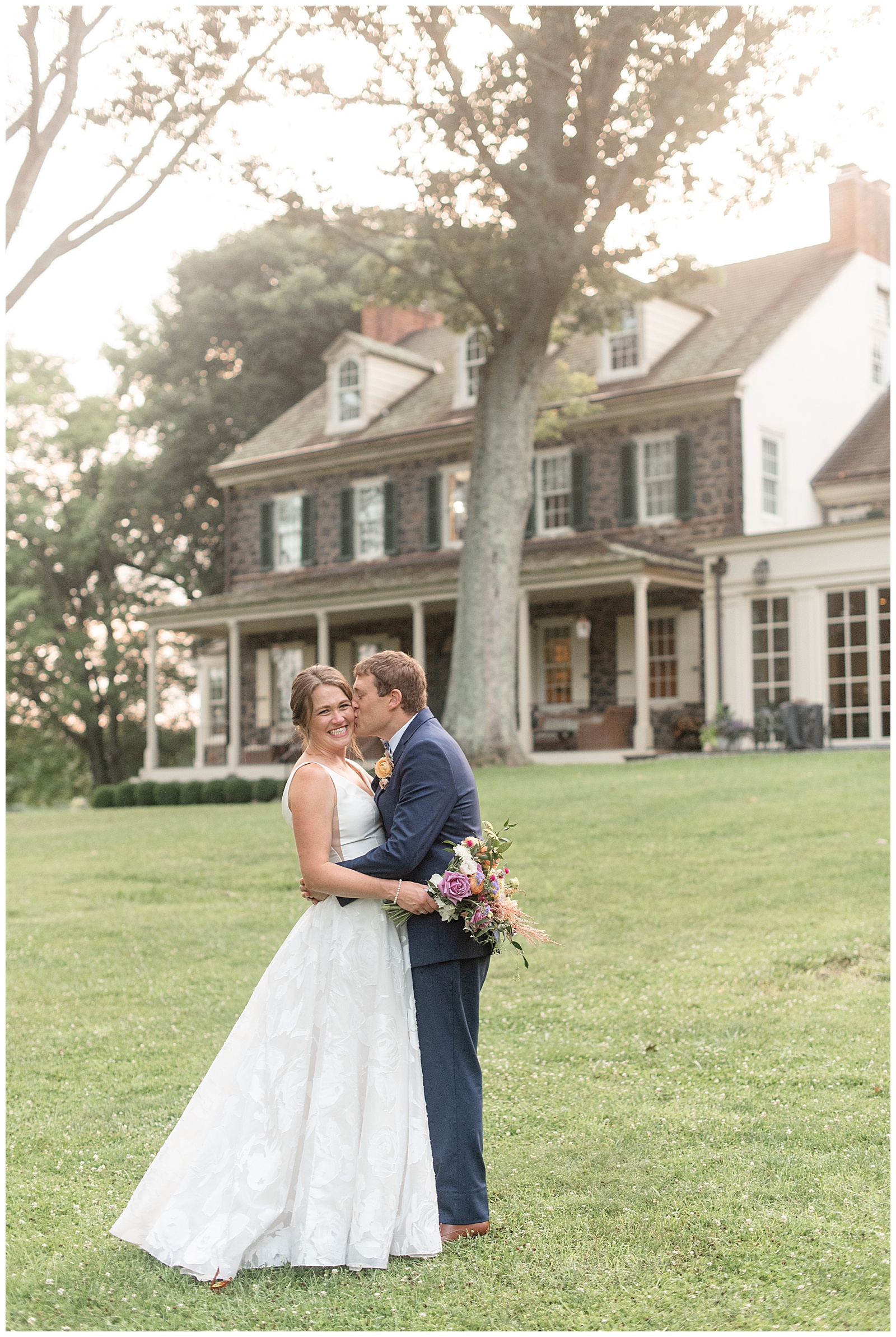 groom in navy blue suit leaning in and kissing bride's left cheek in front of stone home at the inn at grace winery