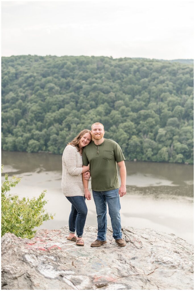 woman resting her left cheek on man's right shoulder as she hugs his right arm on large rock by river at pinnacle overlook in lancaster county