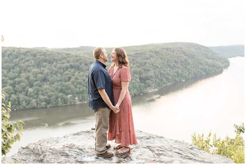 engaged couple standing close holding hands atop large rock overlook the susquehanna river at pinnacle overlook on hazy day
