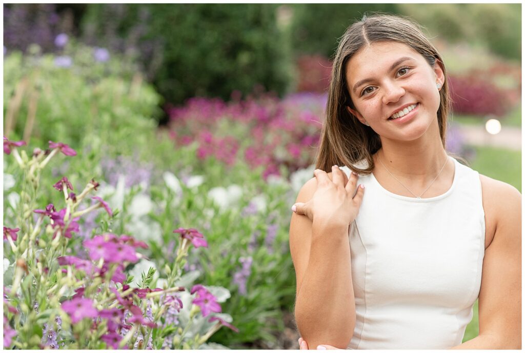 senior girl wearing white tank top with right hand on right shoulder and her head tilted slightly to her right by colorful bush at longwood gardens