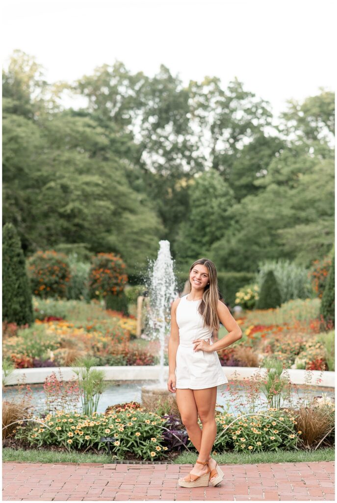 senior girl in white outfit standing in front of fountain with her left hand on her hip and her head tilted left by colorful flowers at longwood gardens