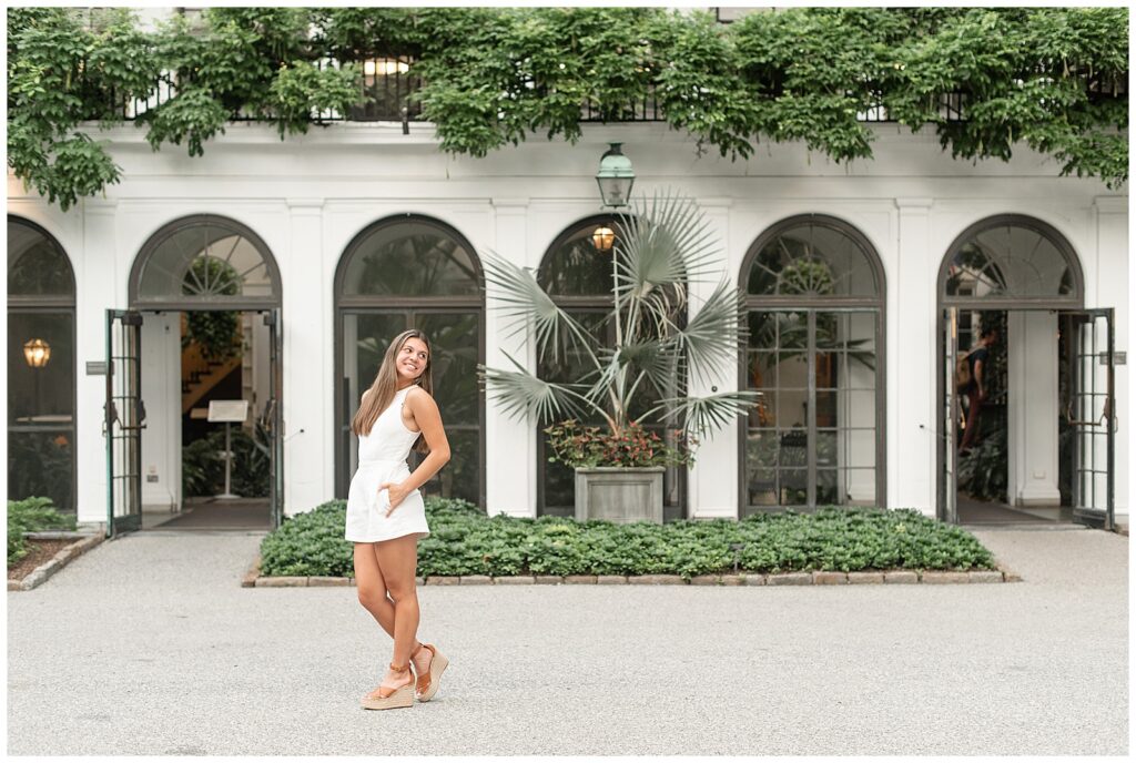 senior girl with hands on hips leaning back looking over her left shoulder in front of white building with lots of archways at longwood gardens