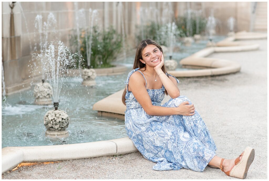 senior girl in light blue and white floral dress sitting on low edge of small fountain at longwood and resting her chin on her left hand