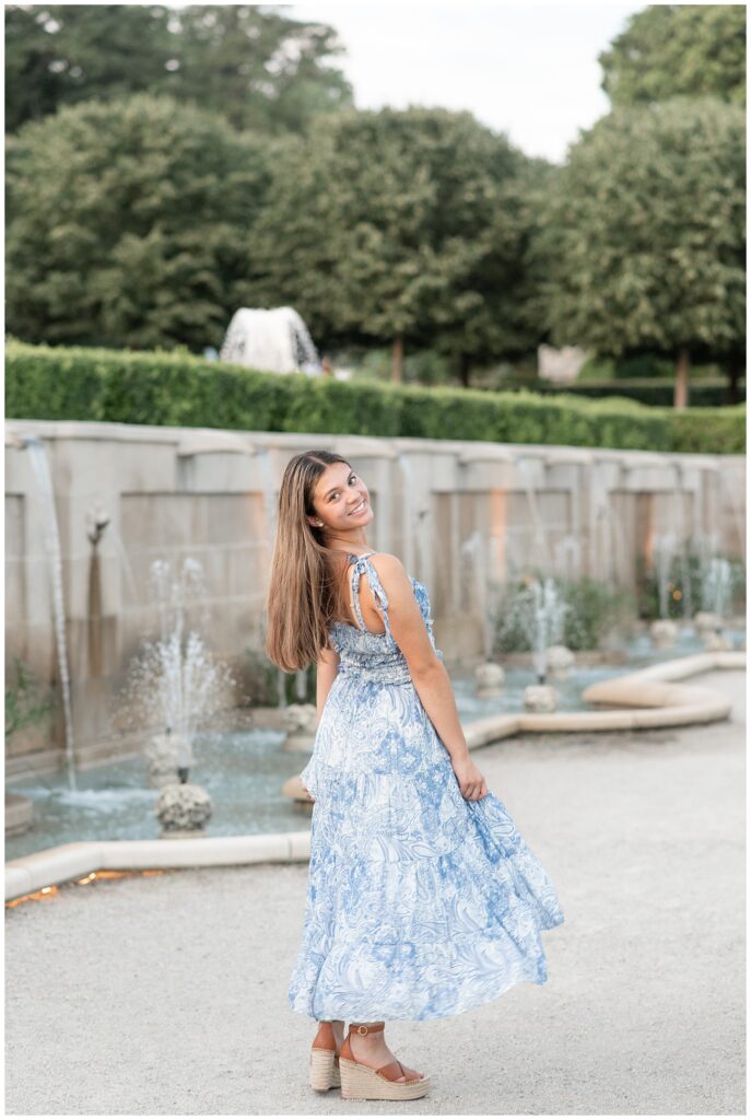 senior girl looking back over right shoulder and holding edges of her dress by small fountains and stone wall at longwood gardens
