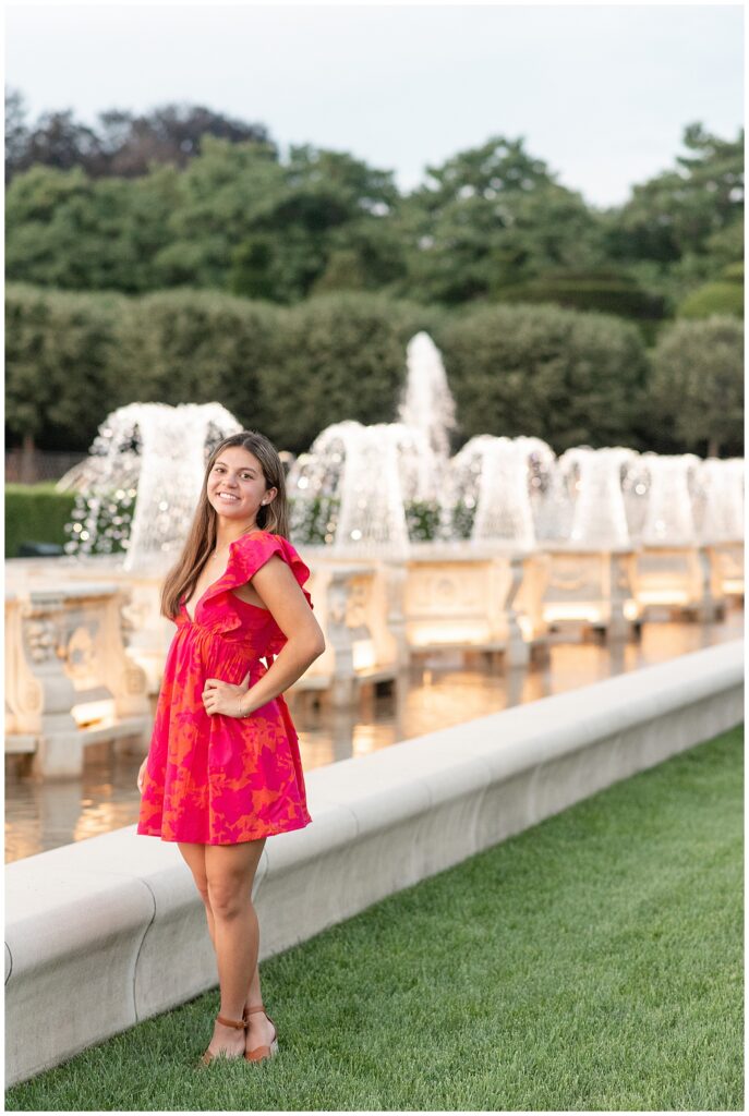 senior girl wearing cute red dress with hands on hips and left shoulder towards camera by row of fountains at longwood gardens