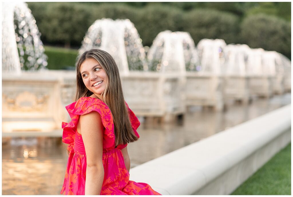 senior girl smiling and looking back over her right shoulder as she sits of ledge of fountains at longwood in kennett square pennsylvania
