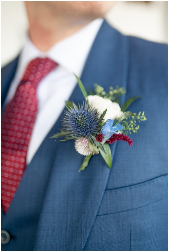 closeup photo of groom's red, white, and blue boutonniere against his navy blue suit and dark red tie at the barn at silverstone