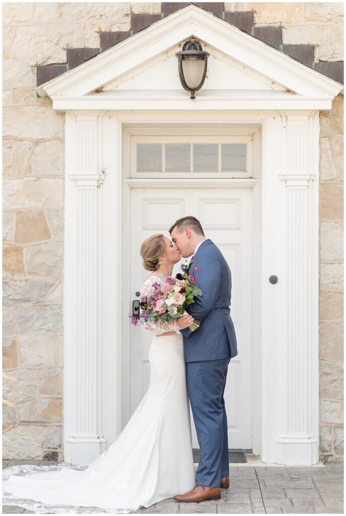 bride and groom hugging and kissing by white doorway of stone building at the barn at silverstone
