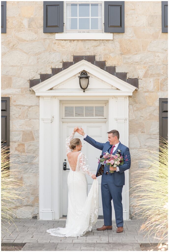 groom twirling his bride under his right hand as he holds her bouquet by white door at the barn at silverstone