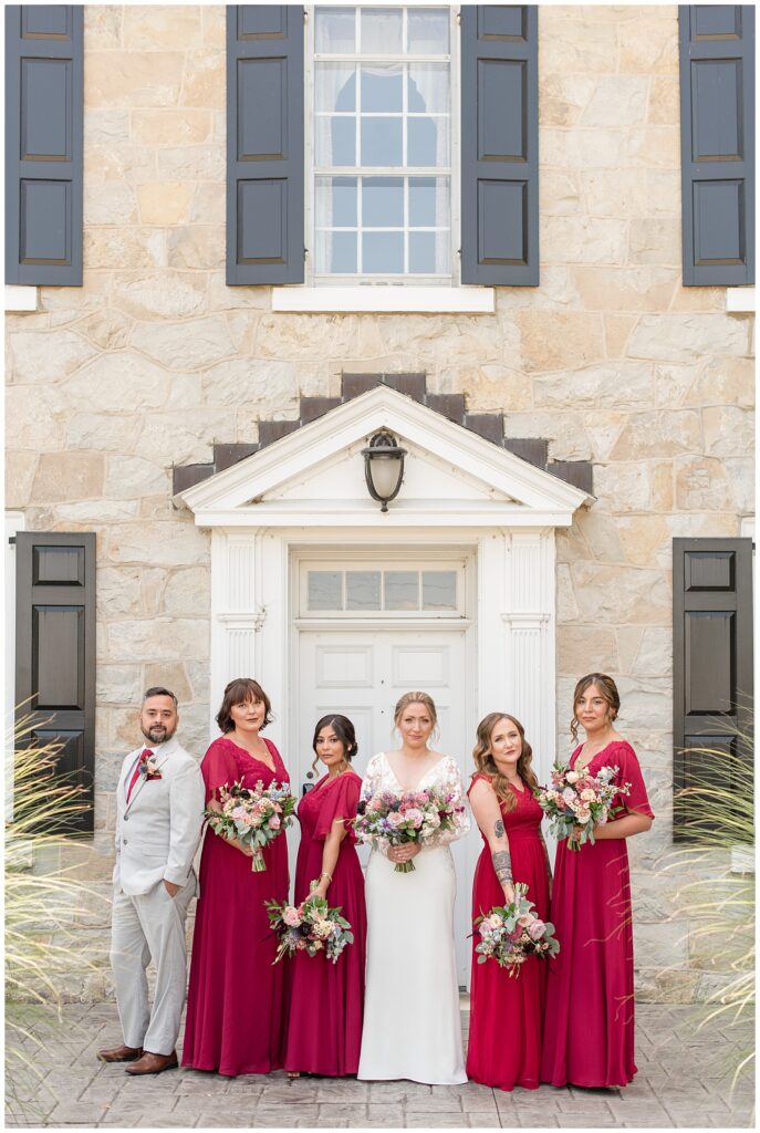 bride surrounded by her bridal party of four girls in red dresses and one guy in tan suit with red tie at the barn at silverstone