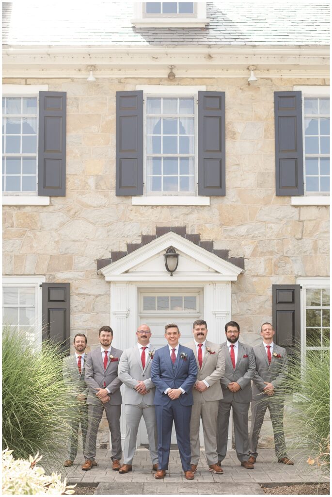 groom surrounded by his five groomsmen wearing shades of gray suits by stone house at the barn at silverstone
