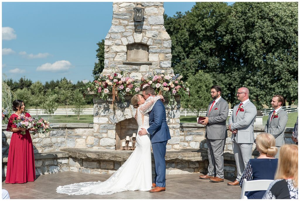couple shares their first kiss during outdoor wedding ceremony by stone fireplace at the barn at silverstone