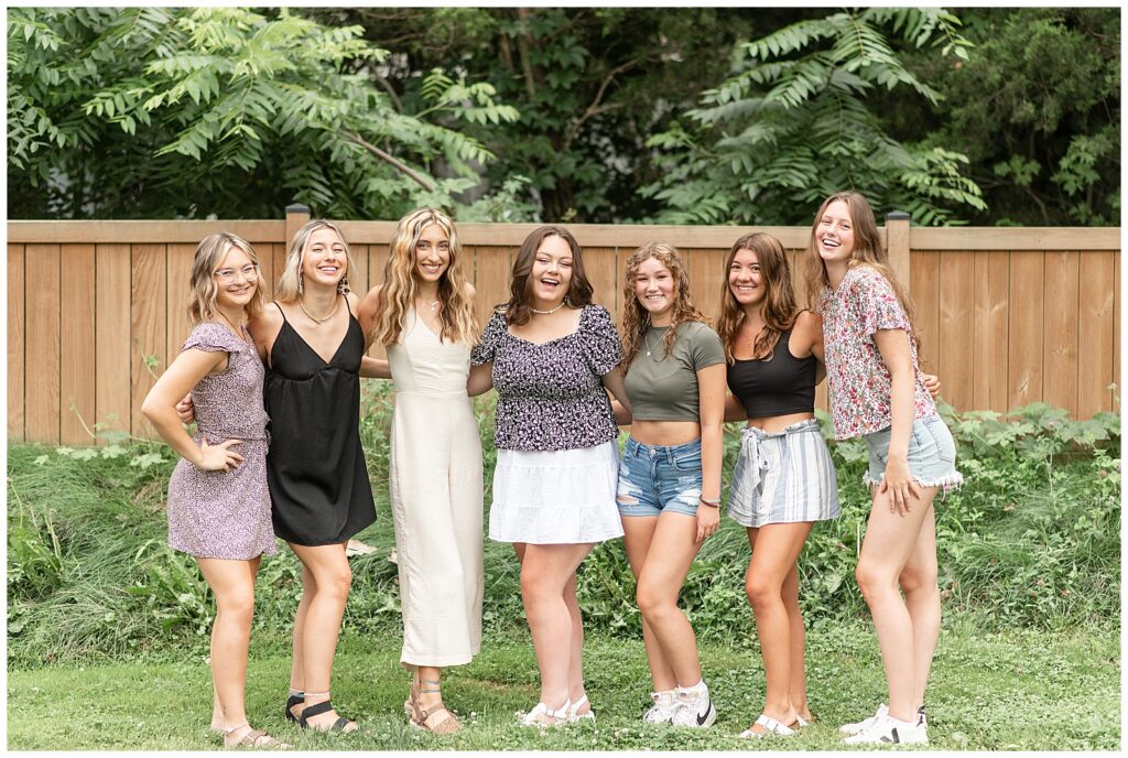 seven senior girl spokesmodels all standing close and leaning in toward one another by wooden fence in lancaster county pennslvania