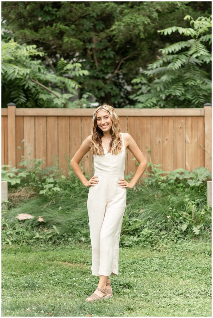 senior girl in white sleeveless romper with both hands on her hips smiling by wooden fence in lancaster pa
