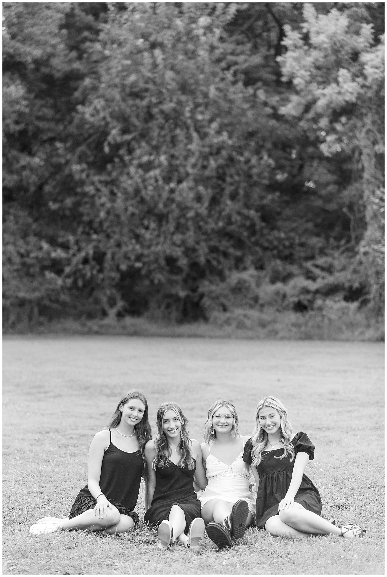 black and white photo of four senior girls all sitting on the grass with trees behind them at park in lancaster county pennsylvania