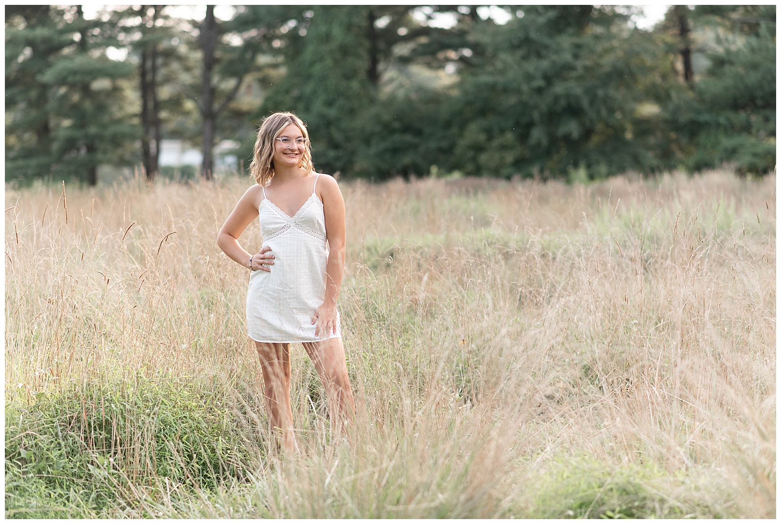 senior girl in white dress standing in field of wild grasses with her right hand on her hip at lancaster county park in pennsylvania