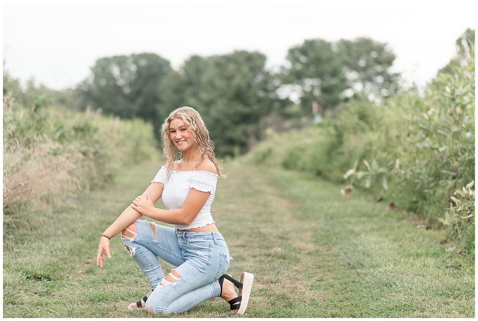 senior girl in white off-the-shoulder top and blue jeans crouched down with hands over knee in grass pathway at park in lancaster county