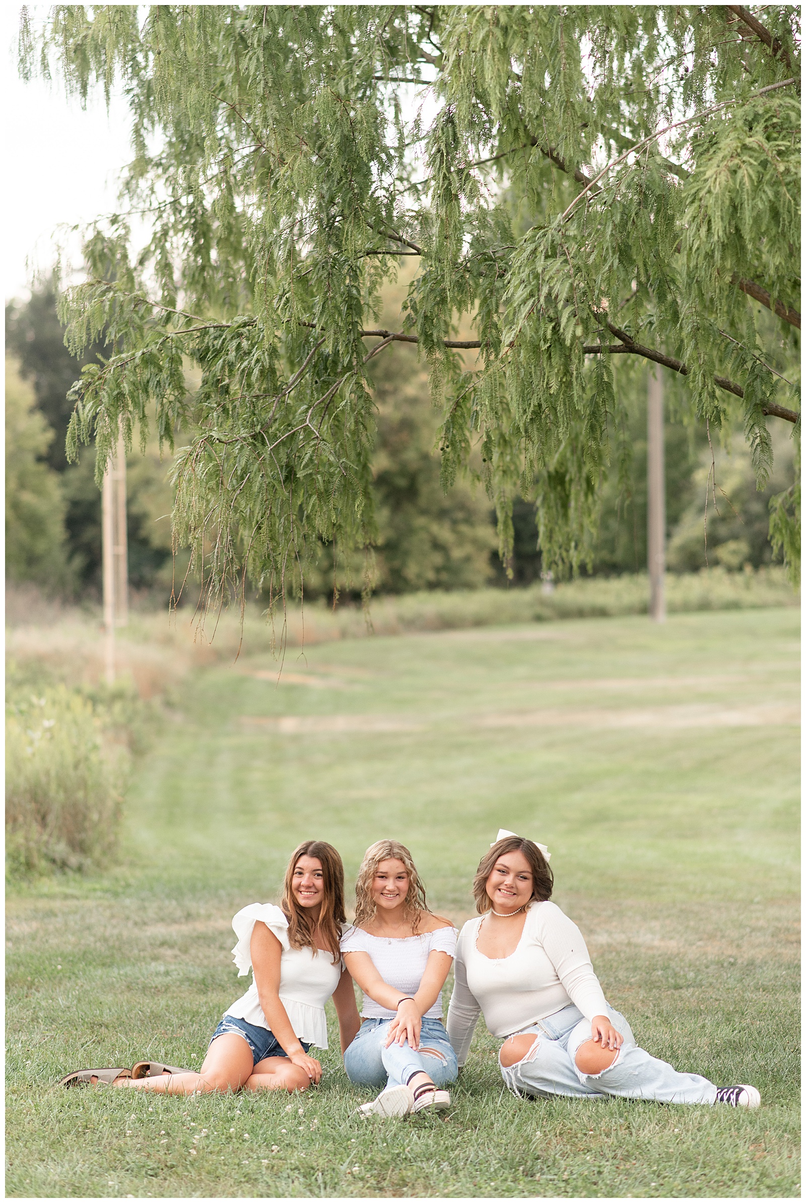 three senior spokesmodel girls in white tops and blue jeans sitting in grass with trees behind them at park in lancaster pennsylvania