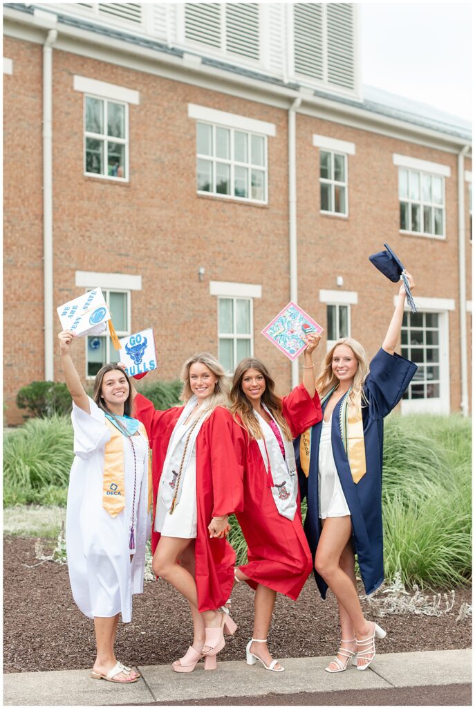 four senior girls in their graduation gowns holding their caps high above them by brick building in hershey pennsylvania