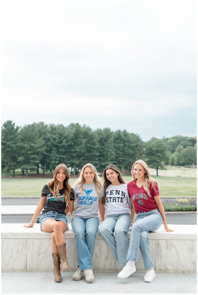 four senior girls sitting along edge of fountain each wearing their college t-shirt and jeans at founders hall