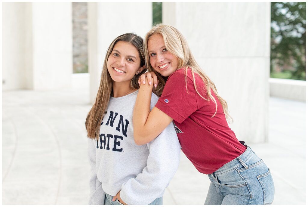 senior girl in red shirt leaning onto left shoulder of other senior girl in penn state sweatshirt at founders hall