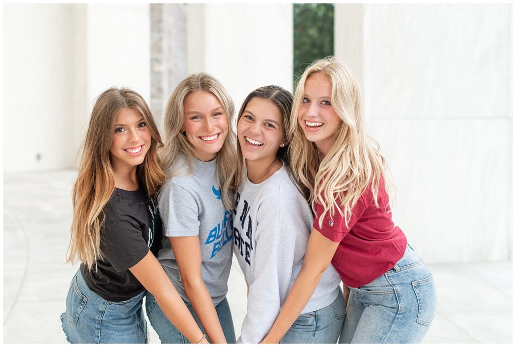 four senior girls huddled together and leaning towards camera hold hands at founders hall