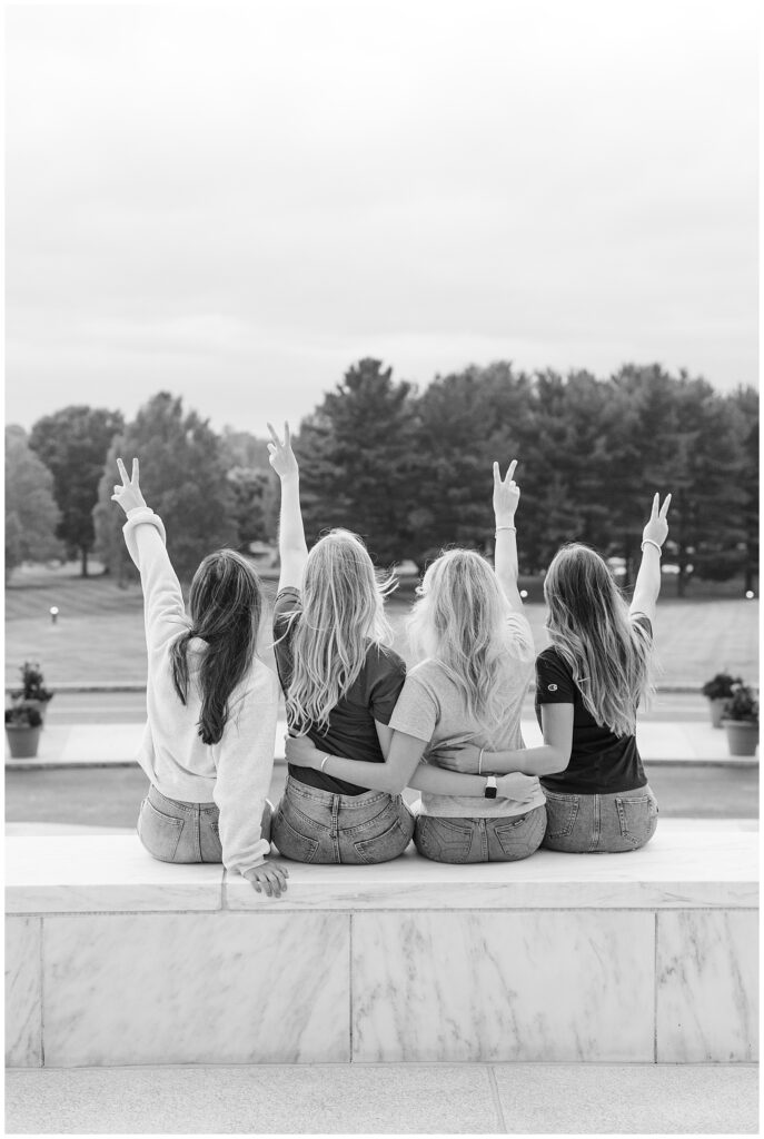 black and white photo of four senior girls sitting on wall with backs toward camera making peace signs in the air in hershey pa