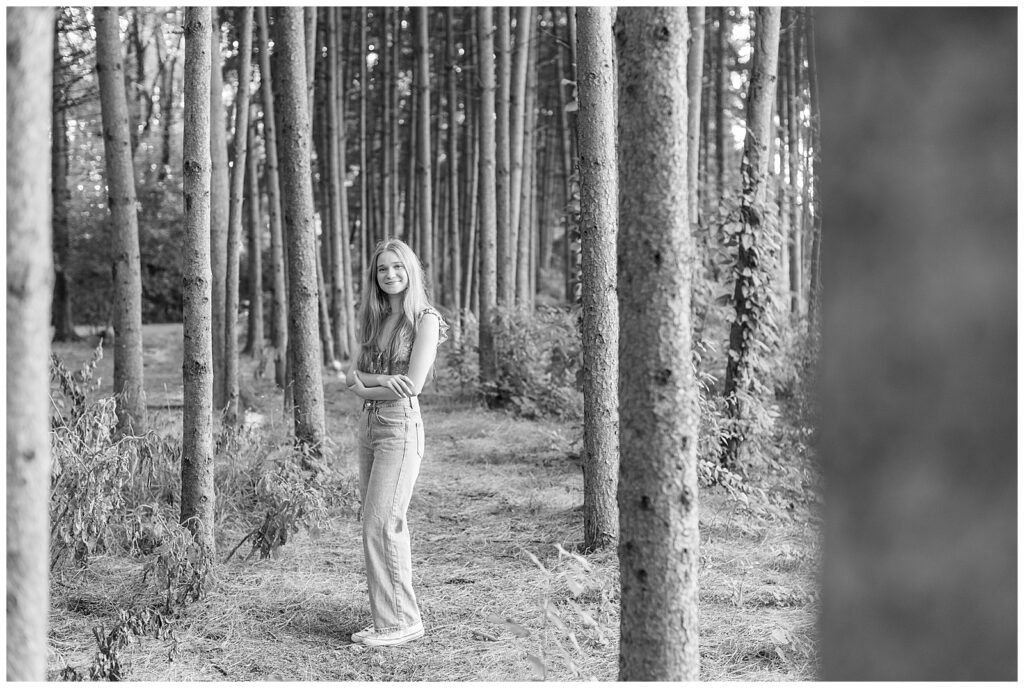 black and white photo of senior girl standing among rows of evergreen trees with left shoulder towards camera at overlook park