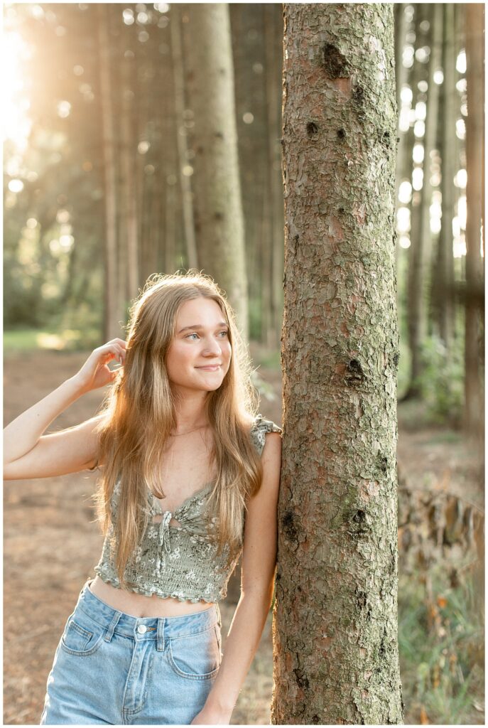 senior girl leaning left shoulder against evergreen tree as she looks left and moves her right hand through her long hair at sunset at overlook park
