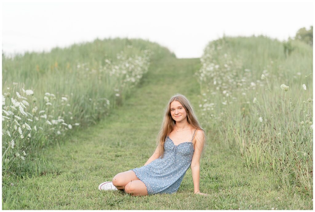 senior girl sitting in grass by white wildflowers and wearing cute blue dress at overlook park in lancaster pa