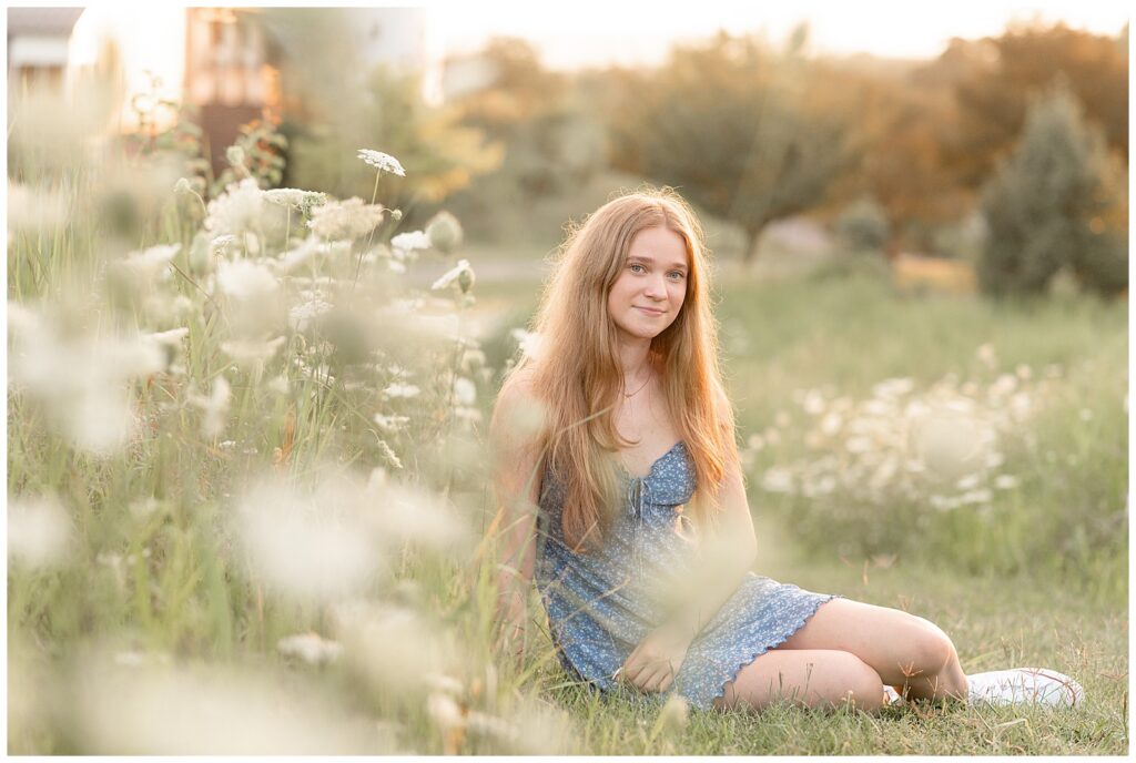 senior girl sitting in white wildflowers slightly smiling at camera at sunset at overlook park