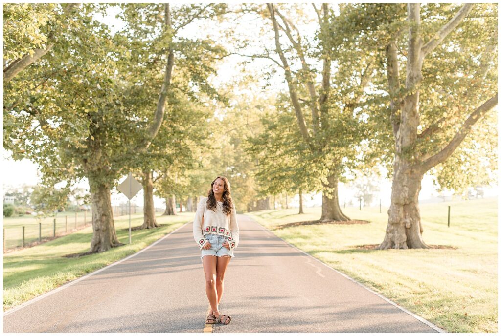 senior girl standing in middle of tree-lined road with hands in her jean short pockets wearing white sweater crop top at masonic village