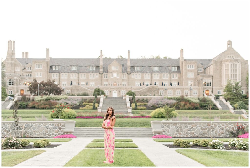 senior girl in pink and white dress standing in beautifully landscaped yard with majestic building behind her at masonic village