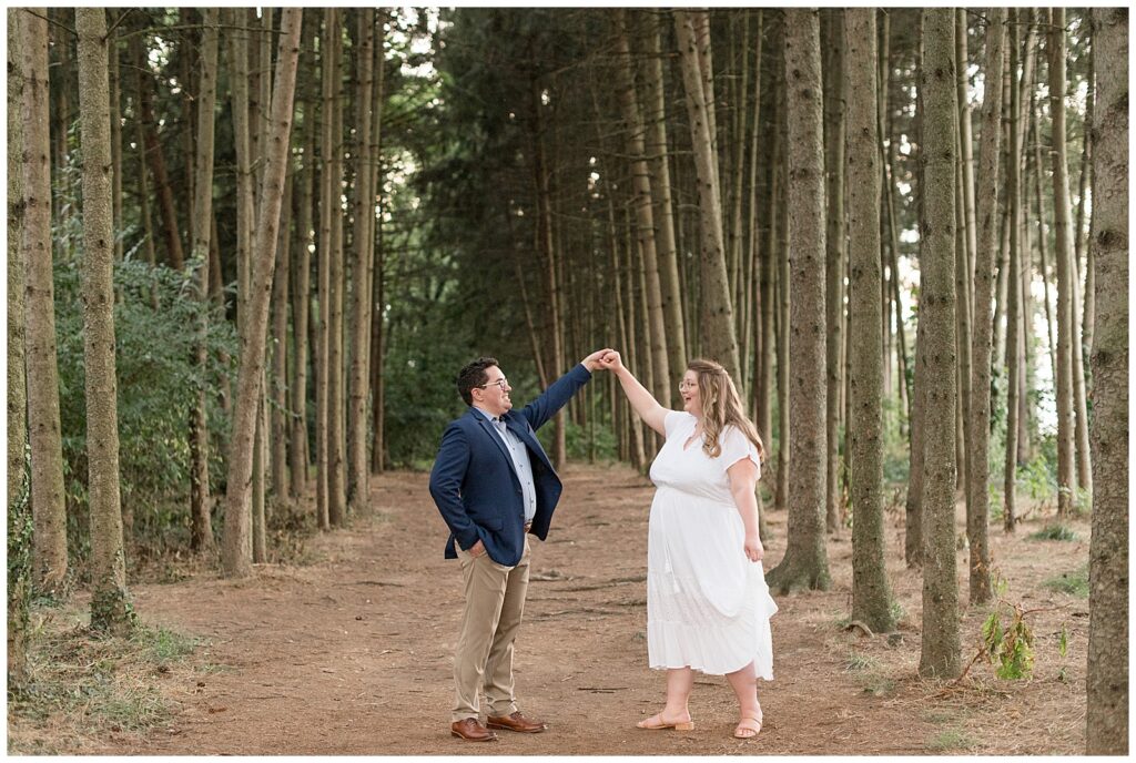 guy in navy suit coat and khaki pants and woman in flowy white dress being twirled under guy's left arm at overlook park by trees