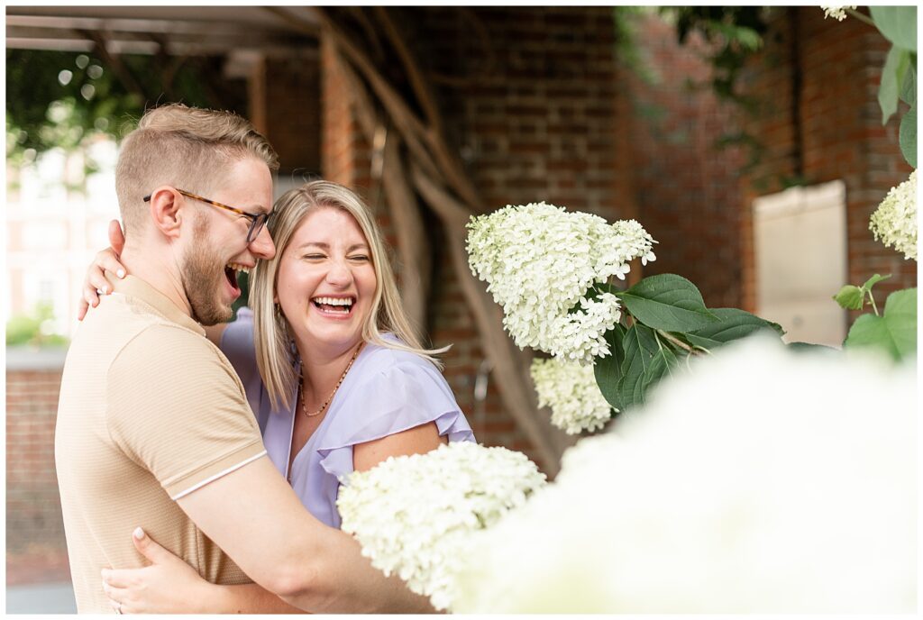 engaged couple laughing as they both look left by white hydrangea bush in philly