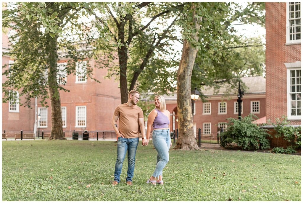 couple holding hands in yard with guy in tan shirt and jeans and girl in brown top and jeans with brick building behind them in philly