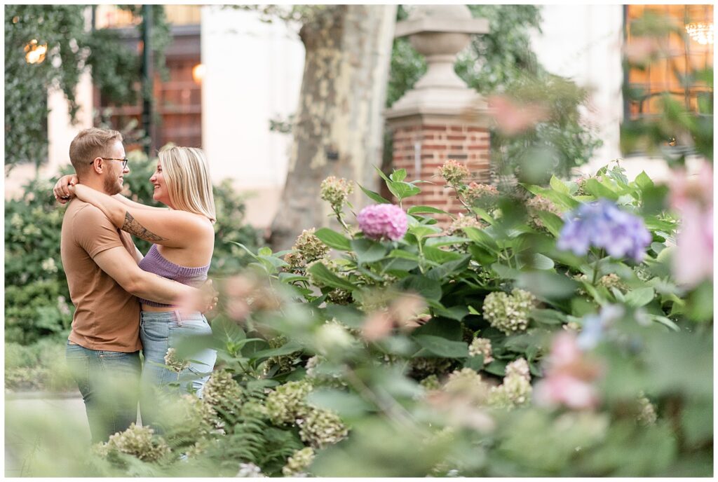 engaged couple hugging and smiling at each other behind colorful hydrangea bushes in philly