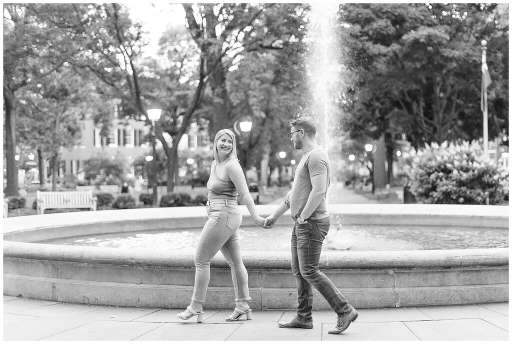 black and white photo of girl holding guy's hand and leading as she looks back by fountain in philadelphia, pa