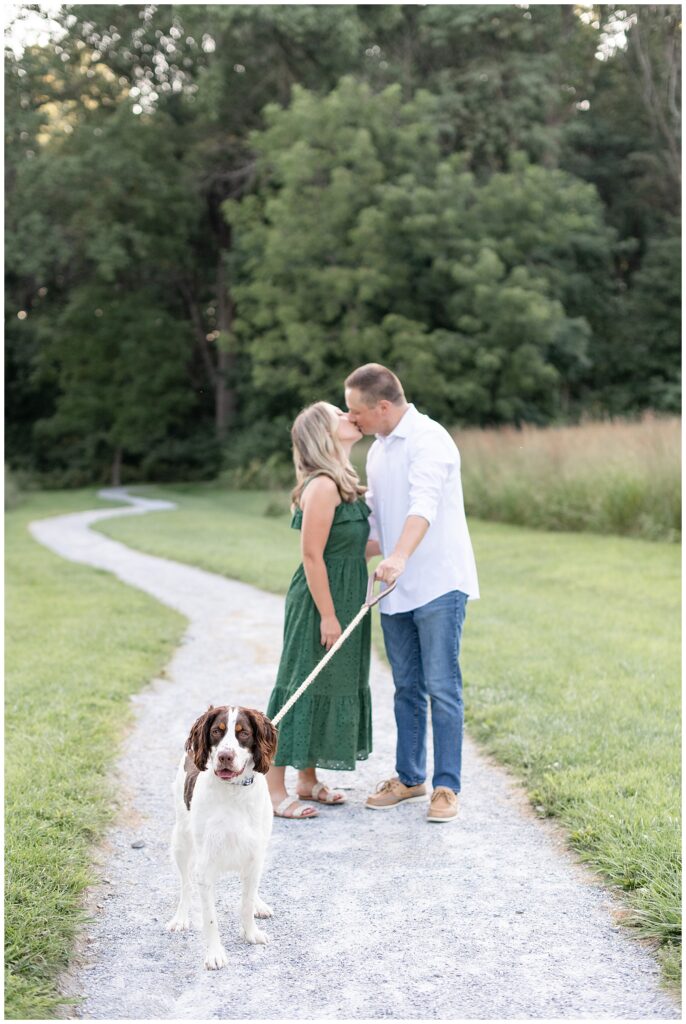 Couple kissing on stone pathway as guy in light dress shirt and jeans holds leash of their dog looking at the camera in front of them and girl wears long dark green dress at hibernia park