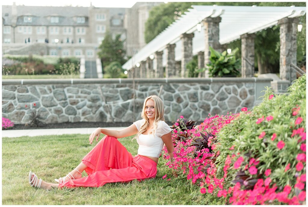 senior girl sitting in lawn wearing pink long skirt and white t-shirt by stone wall at pink bushes at masonic village