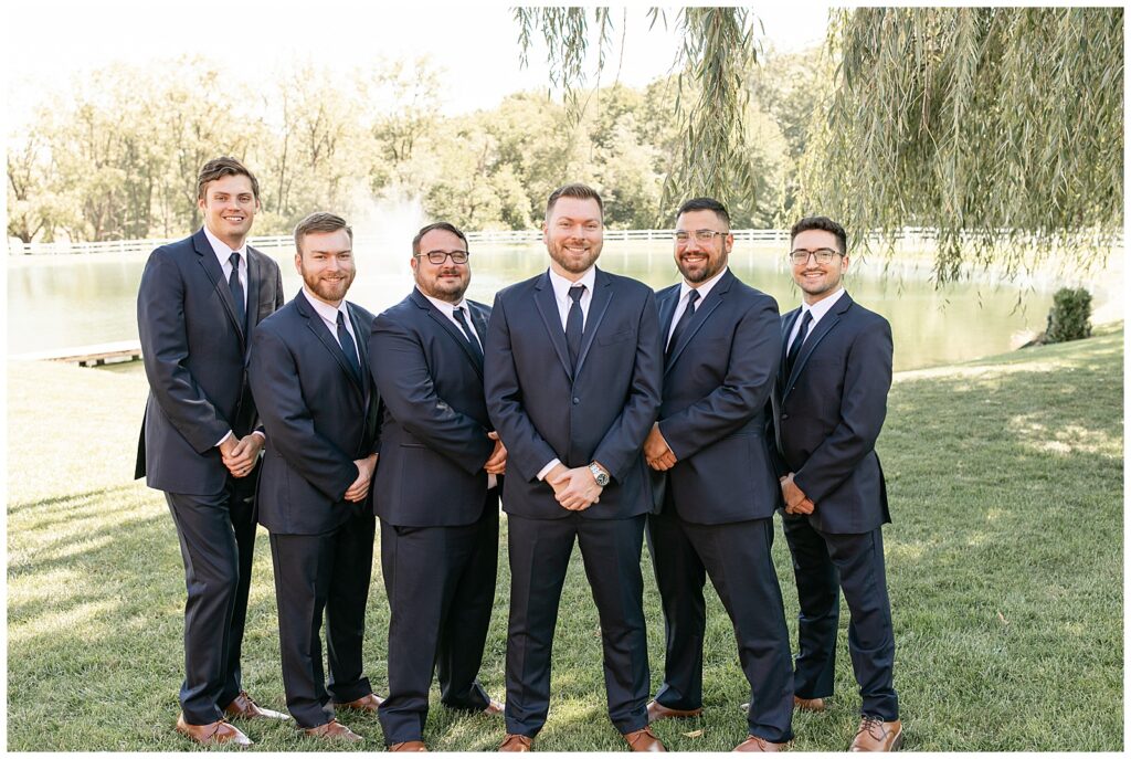 groom surrounded by his five groomsmen all in dark suits and hands folded in front of them by pond at pond view farm
