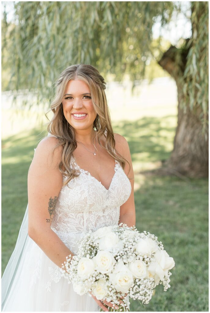 bride smiling at camera in sleeveless v-neck white wedding gown and holding bouquet of white flowers at pond view farm